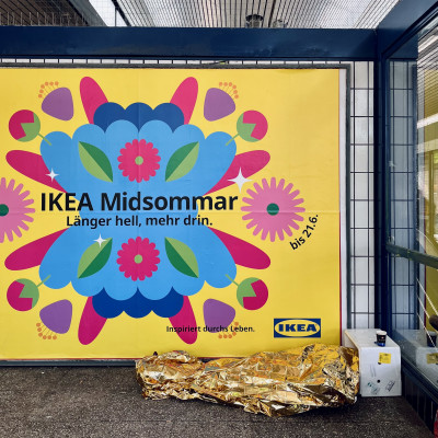 Person mit goldener Rettungsdecke vor einem IKEA-Werbeplakat im öffentlichen Raum